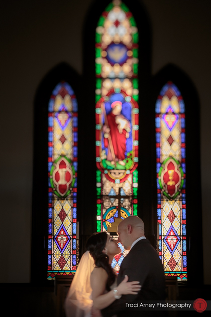 bride and groom share kiss in front of intricate stained glass windows in St. Athanasius Church, Burlington, NC